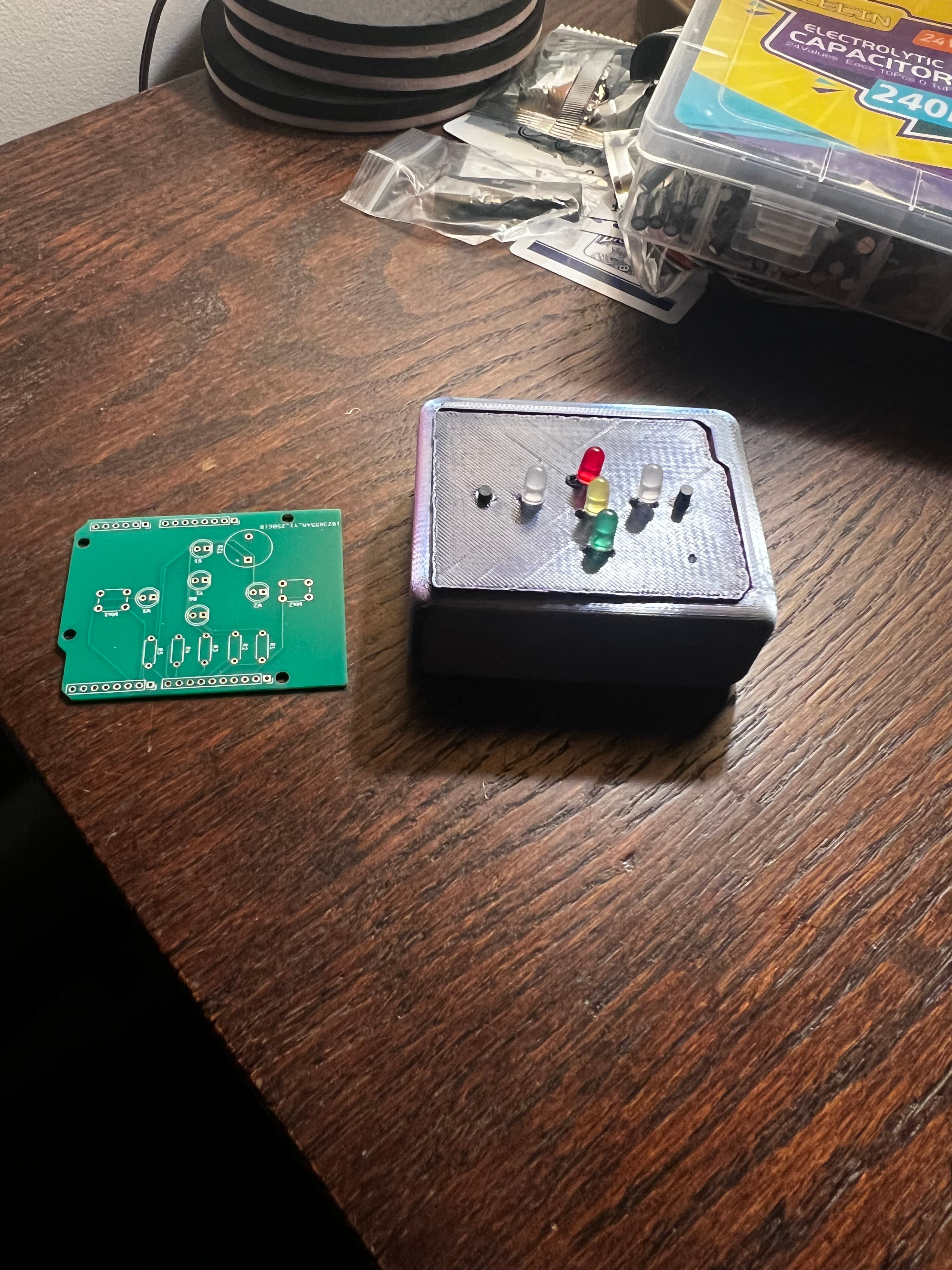 Bare green PCB next to 3D printed enclosure with LEDs poking through lid, sitting on desk